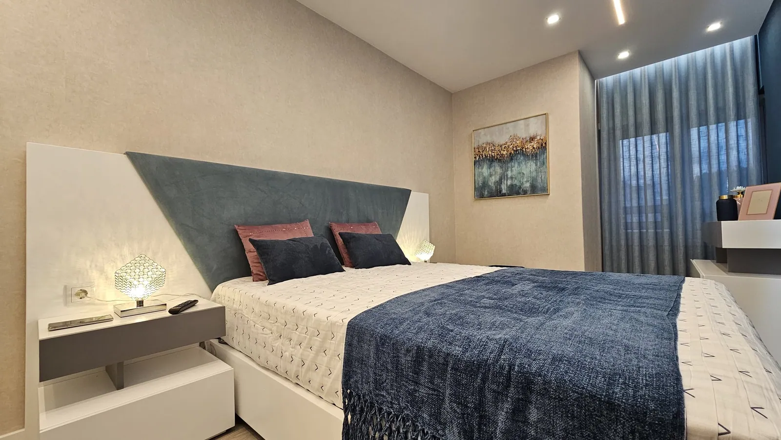 O quarto com a cabeceira estofada azul em diagonal e as cortinas.