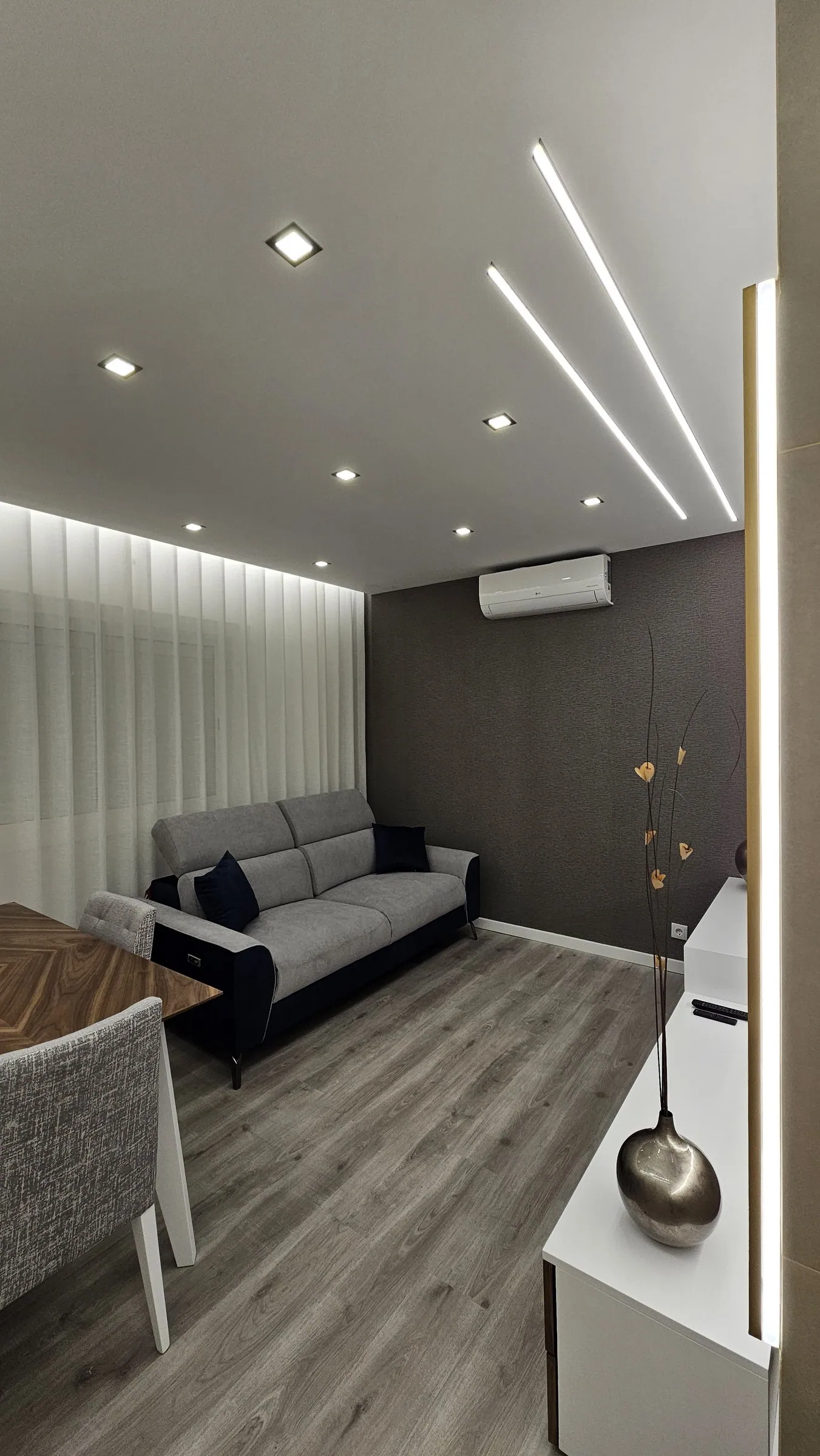 Sala com sofá cinzento, cortinados claros e luz linear no teto.