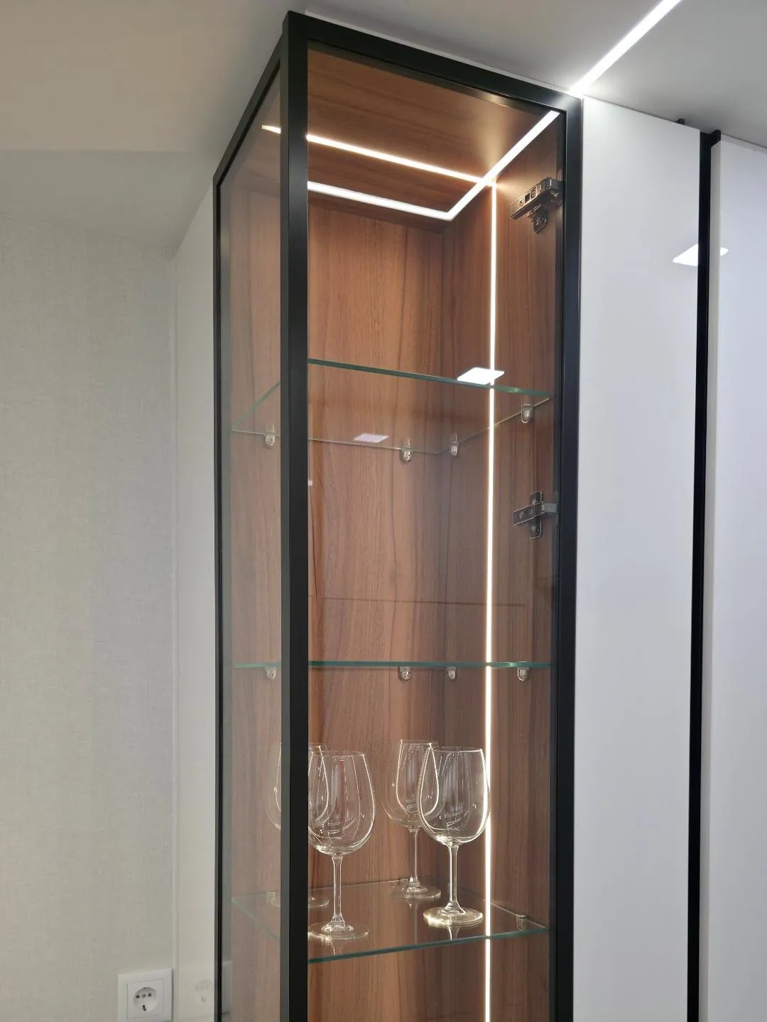 Vitrine de vidro em caixilho preto com copos e madeira iluminada.